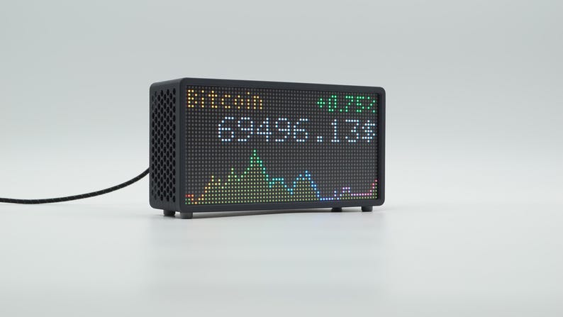 Peut inclure: Un &eacute;cran d'affichage num&eacute;rique rectangulaire noir avec un motif lat&eacute;ral en nid d'abeille. L'&eacute;cran affiche le texte Bitcoin, la valeur 69496.13$, et un indicateur vert plus 0.75% au-dessus d'un graphique lin&eacute;aire pix&eacute;lis&eacute; color&eacute;. Un c&acirc;ble noir d&eacute;passe.