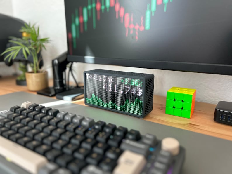 Peut inclure: Un &eacute;cran num&eacute;rique rectangulaire noir affichant le texte Tesla Inc. et le nombre 411.74$ avec un graphique lin&eacute;aire vert en dessous. L'appareil pr&eacute;sente un &eacute;cran &agrave; matrice de points et un bo&icirc;tier textur&eacute;, pos&eacute; sur un bureau en bois.
