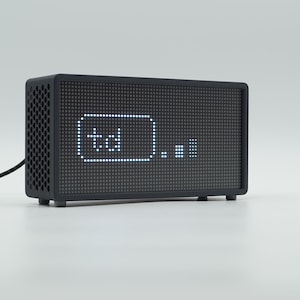 Peut inclure: Une horloge num&eacute;rique rectangulaire noire avec un &eacute;cran &agrave; matrice de points affichant le texte td et une petite ic&ocirc;ne de graphique &agrave; barres. L'appareil pr&eacute;sente un panneau lat&eacute;ral perfor&eacute;, un cordon d'alimentation noir et de petits pieds.