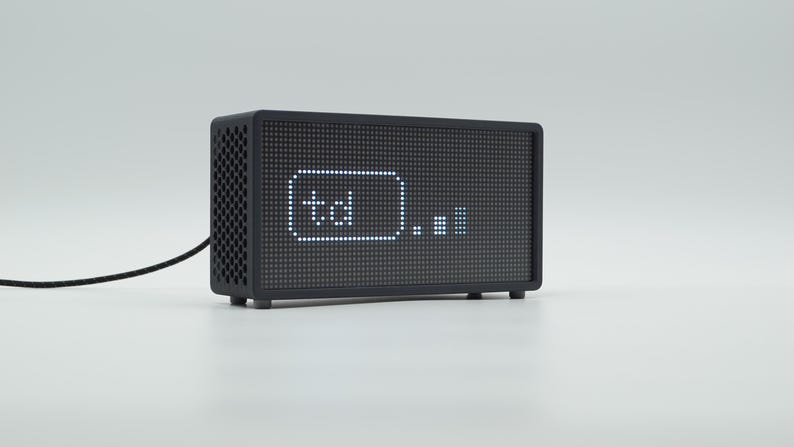 Peut inclure: Une horloge num&eacute;rique rectangulaire noire avec un &eacute;cran &agrave; matrice de points affichant le texte td et une petite ic&ocirc;ne de graphique &agrave; barres. L'appareil pr&eacute;sente un panneau lat&eacute;ral perfor&eacute;, un cordon d'alimentation noir et de petits pieds.