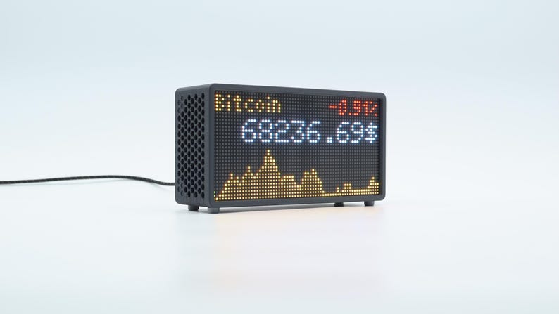 May include: A black digital display showing Bitcoin data. The screen displays "Bitcoin" in yellow, a negative percentage in red, and the current price in white. A yellow and orange graph is at the bottom.