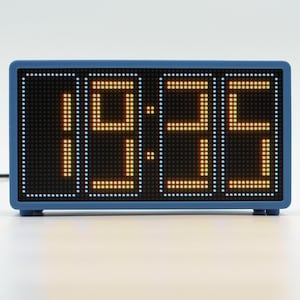 Peut inclure: Une horloge num&eacute;rique rectangulaire avec un cadre bleu et un &eacute;cran noir affichant l'heure 19:35 en chiffres LED orange &agrave; matrice de points. Un fin cordon d'alimentation noir s'&eacute;tend sur le c&ocirc;t&eacute; gauche de l'appareil pos&eacute; sur une surface.