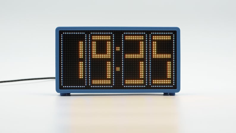 Peut inclure: Une horloge num&eacute;rique rectangulaire avec un cadre bleu et un &eacute;cran noir affichant l'heure 19:35 en chiffres LED orange &agrave; matrice de points. Un fin cordon d'alimentation noir s'&eacute;tend sur le c&ocirc;t&eacute; gauche de l'appareil pos&eacute; sur une surface.