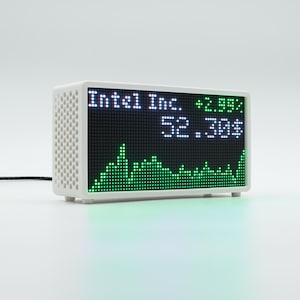 Peut inclure: Un appareil d'affichage num&eacute;rique blanc rectangulaire avec un panneau lat&eacute;ral perfor&eacute;. L'&eacute;cran affiche le texte Intel Inc., la valeur 52.30$, un pourcentage de +2.99% et un graphique lin&eacute;aire vert repr&eacute;sentant des donn&eacute;es boursi&egrave;res. Un fin c&acirc;ble noir est attach&eacute;.