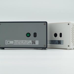 Peut inclure: Deux appareils &eacute;lectroniques rectangulaires, l'un avec une fa&ccedil;ade gris fonc&eacute; et l'autre avec un bo&icirc;tier blanc. L'appareil gris pr&eacute;sente un bouton circulaire vert, un port USB-C et une &eacute;tiquette avec le texte td, mod&egrave;le TIC05 1 et un QR code.