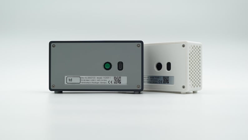 Peut inclure: Deux appareils &eacute;lectroniques rectangulaires, l'un avec une fa&ccedil;ade gris fonc&eacute; et l'autre avec un bo&icirc;tier blanc. L'appareil gris pr&eacute;sente un bouton circulaire vert, un port USB-C et une &eacute;tiquette avec le texte td, mod&egrave;le TIC05 1 et un QR code.