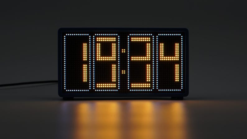 Peut inclure: Une horloge num&eacute;rique rectangulaire avec un cadre sombre affichant l'heure 19:34 en chiffres orange de style matrice de points. L'horloge pr&eacute;sente une bordure blanche de petits points lumineux entourant l'affichage central orange sur &eacute;cran noir.