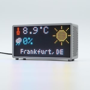 Peut inclure: Un affichage m&eacute;t&eacute;o num&eacute;rique rectangulaire avec un bo&icirc;tier gris et un panneau lat&eacute;ral perfor&eacute;. L'&eacute;cran affiche une ic&ocirc;ne de thermom&egrave;tre rouge, 8,9 C, une ic&ocirc;ne de soleil jaune, une ic&ocirc;ne de nuage bleu avec 0% et le texte Frankfurt, DE en blanc.