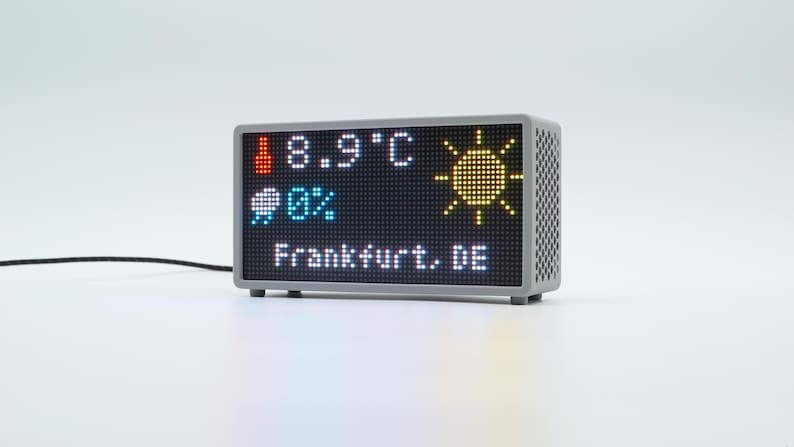 Peut inclure: Un affichage m&eacute;t&eacute;o num&eacute;rique rectangulaire avec un bo&icirc;tier gris et un panneau lat&eacute;ral perfor&eacute;. L'&eacute;cran affiche une ic&ocirc;ne de thermom&egrave;tre rouge, 8,9 C, une ic&ocirc;ne de soleil jaune, une ic&ocirc;ne de nuage bleu avec 0% et le texte Frankfurt, DE en blanc.