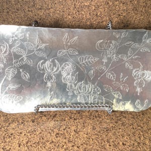 Könnte beinhalten: Ein rechteckiges Tablett aus silberfarbenem Metall mit gehämmertem Finish und abgerundeten Ecken. Die Oberfläche zeigt ein eingraviertes Design von Fuchsienblüten mit Blättern und Stängeln. Das Tablett steht auf einem gedrehten Metallständer.