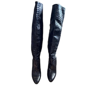 Op de afbeelding: Een paar zwarte kniehoge laarzen met een slangenprint. De laarzen hebben een spitse neus en een hoge hak. De leren laarzen worden tegen een witte achtergrond getoond.