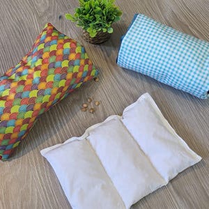 Puede incluir: Tres almohadas rectangulares de aromaterapia. Una tiene un diseño de arcoíris, otra es blanca y la tercera tiene un diseño de cuadros vichy azul claro. También hay una pequeña planta en maceta.
