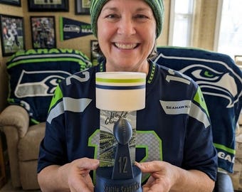Może przedstawiać: Granatowo-zielone trofeum w stylu Seattle Seahawks z nadrukiem "World Champs" i "12". Trofeum trzyma osoba ubrana w koszulkę i czapkę Seahawks.