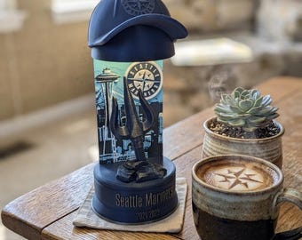 May include: A navy blue Seattle Mariners baseball cap sits atop a decorative item featuring the Seattle skyline, a trident, and the words "Seattle Mariners 2021/2022." A cup of coffee with latte art and a small succulent plant are also visible.
