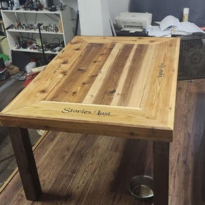 May include: A rectangular wooden table with dark brown legs and a light brown tabletop. The tabletop has a framed edge with the words "Stories Last" and "Legends Rise" engraved on it. The table is in a room with a printer and fan.