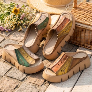 Puede incluir: Sandalias de plataforma de cuero con punta abierta. Las sandalias presentan un diseño multicolor con detalles en verde, amarillo y marrón. Las suelas son de color marrón claro. Las sandalias se exhiben al aire libre sobre una superficie de piedra.