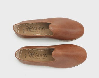 May include: Two brown leather slip-on shoes. The shoes have a rounded toe and a low profile. The brand name "ADEA" and "Handmade in Türkiye" are printed on the insole. The shoes are a size W 38.