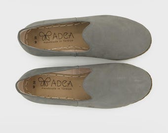 May include: A pair of grey slip-on shoes with a rounded toe. The interior features a tan insole with the text ADEA, a butterfly logo, and the words Handmade in Türkiye. The size W 39 is printed on the side of the insole with visible stitching.