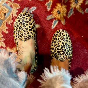 May include: A pair of leopard print slippers with a tan and black spotted pattern. The slippers are on a red patterned rug. The image is a top-down view.