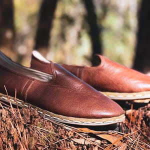 May include: A pair of brown leather slip-on shoes with a stitched sole. The shoes are displayed on a tree stump with a blurred natural background. The shoes have a minimalist design.