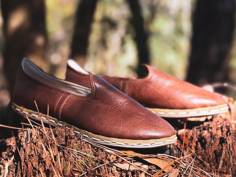 May include: A pair of brown leather slip-on shoes with a stitched sole. The shoes are displayed on a tree stump with a blurred natural background. The shoes have a minimalist design.