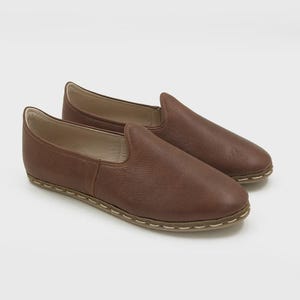 May include: A pair of brown leather slip-on shoes. The shoes have a rounded toe and a low profile. The soles are a lighter color and are stitched to the upper. The shoes are made of a soft, textured leather.
