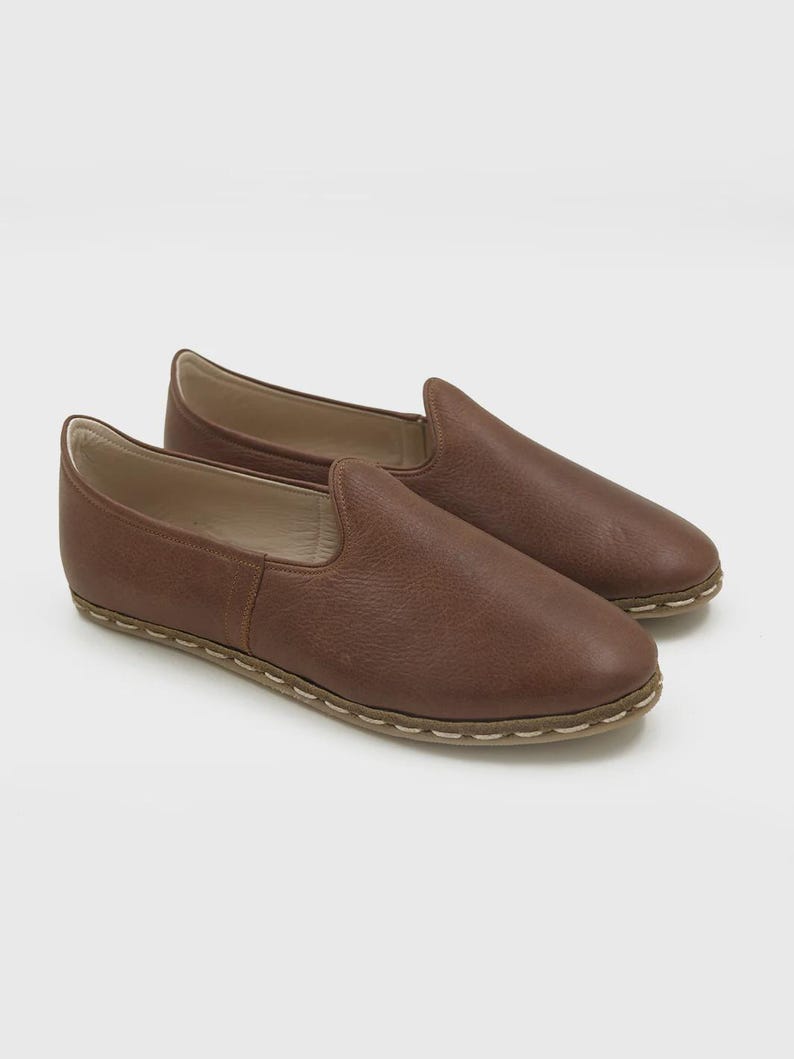 May include: A pair of brown leather slip-on shoes. The shoes have a rounded toe and a low profile. The soles are a lighter color and are stitched to the upper. The shoes are made of a soft, textured leather.