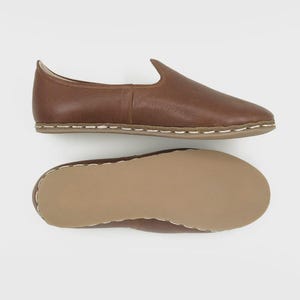 May include: A pair of brown leather slip-on shoes. The shoes have a rounded toe and a low profile. The soles are a light brown color with white stitching around the edges. The top shoe is shown from the side, and the bottom shoe shows the sole.
