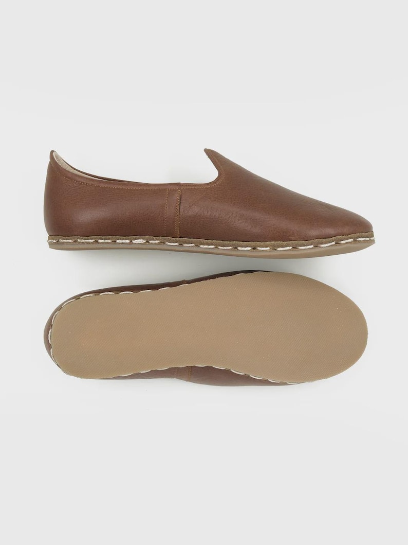 May include: A pair of brown leather slip-on shoes. The shoes have a rounded toe and a low profile. The soles are a light brown color with white stitching around the edges. The top shoe is shown from the side, and the bottom shoe shows the sole.