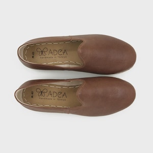 May include: A pair of brown leather slip-on shoes. The shoes have a rounded toe and a stitched sole. The insole is stamped with the brand name "ADEA Handmade in T&uuml;rkiye". The shoes are size M 43.
