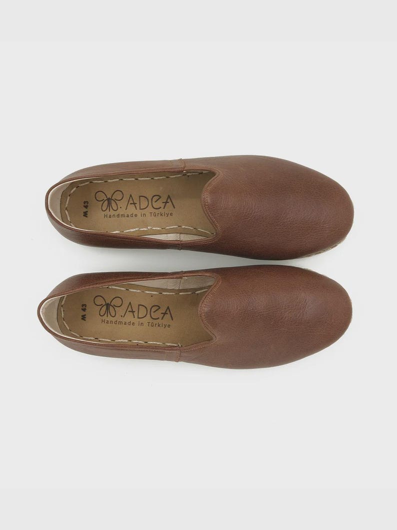 May include: A pair of brown leather slip-on shoes. The shoes have a rounded toe and a stitched sole. The insole is stamped with the brand name "ADEA Handmade in T&uuml;rkiye". The shoes are size M 43.