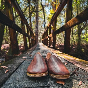 May include: A pair of brown leather slip-on shoes with a stitched sole, resting on a wooden bridge. The shoes are the focal point, with a blurred background of a forest and a wooden bridge railing. The shoes appear to be handmade.