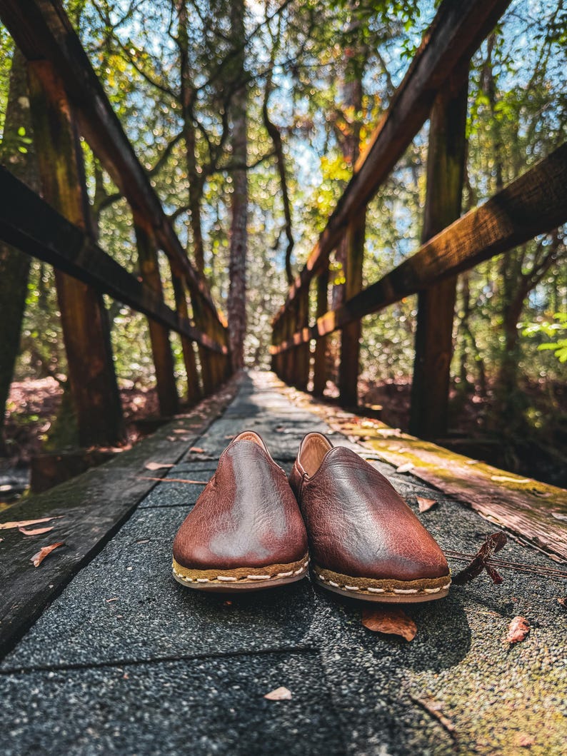 May include: A pair of brown leather slip-on shoes with a stitched sole, resting on a wooden bridge. The shoes are the focal point, with a blurred background of a forest and a wooden bridge railing. The shoes appear to be handmade.
