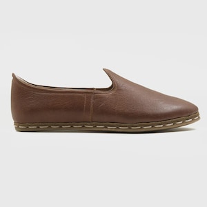 May include: A brown leather slip-on shoe with a pointed toe. The shoe has a textured surface and a stitched sole. The overall design is minimalist and casual, suitable for everyday wear. The shoe's color is a warm, earthy brown.
