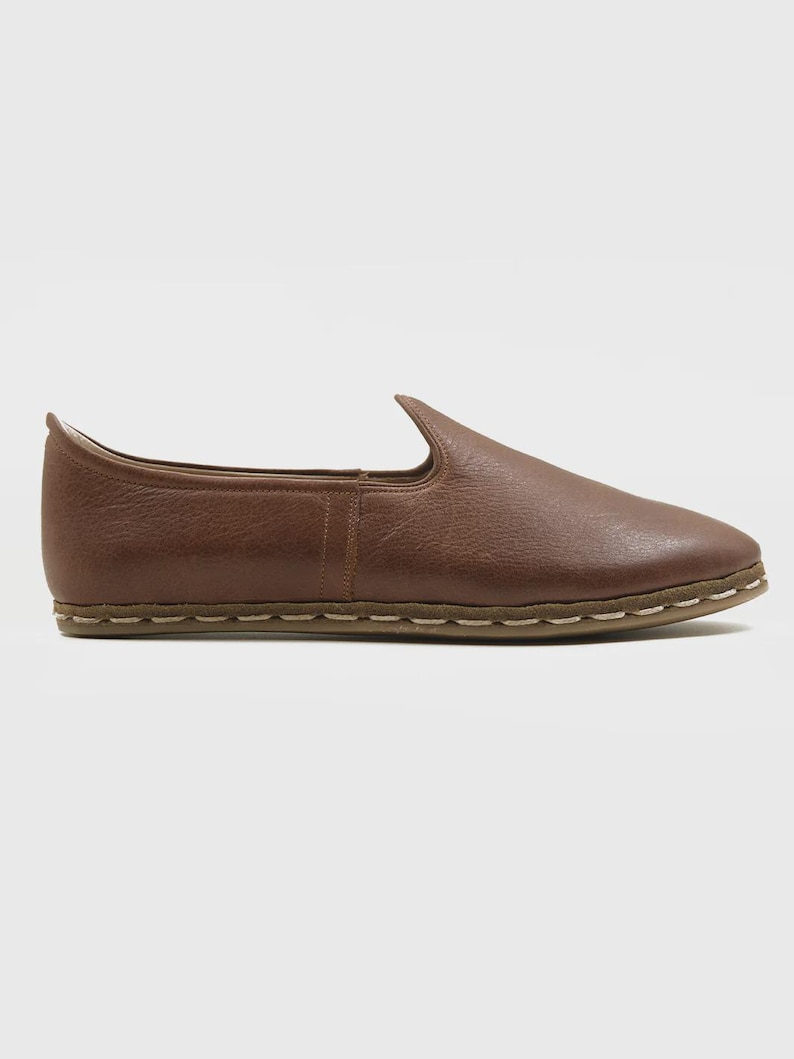 May include: A brown leather slip-on shoe with a pointed toe. The shoe has a textured surface and a stitched sole. The overall design is minimalist and casual, suitable for everyday wear. The shoe's color is a warm, earthy brown.