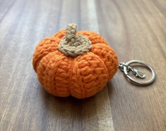 May include: An orange crocheted pumpkin keychain with a tan stem. The pumpkin is round with ribbed detailing. The keychain has a silver-colored ring and clasp.
