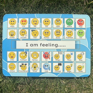 May include: A rectangular blue communication board featuring a grid of yellow emoji faces representing various emotions and states. The centre text reads I am feeling followed by five dots. Each emoji is labelled with words like Happy, Love, and Hungry.