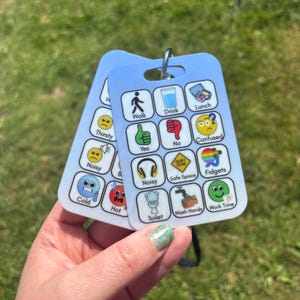 May include: A light blue rectangular communication card featuring a grid of icons with labels including Walk, Drink, Lunch, Yes, No, Confused, Noisy, Safe Space, Fidgets, Cold, Hot, Toilet, Wash Hands, and Work Time, attached to a metal ring.