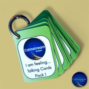 May include: A set of green and white "I am feeling... Talking Cards Pack 1" attached to a silver metal ring. The cards have a blue and white logo that says "Calmstream Studio". The background is a light yellow colour.