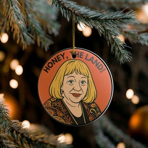 May include: Circular ceramic Christmas ornament with a cartoon illustration of a woman with blonde hair and the text "HONEY, THE LAND!". The ornament has a gold hanging string and is set against a blurred Christmas tree background with lights.