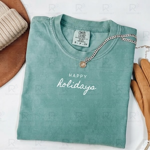 May include: A teal Comfort Colors t-shirt with the words "HAPPY holidays" in white script. A silver chain necklace with a pendant and a pair of brown leather gloves are also visible.