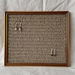 Può includere: Una cornice rettangolare color oro contenente un pannello in tessuto beige lavorato all'uncinetto. Due paia di orecchini pendenti color argento con piccole perline bianche sono fissati sulla superficie tessuta e ruvida del pannello.
