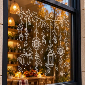 May include: A white line art window decal featuring hanging autumn elements including sunflowers, pumpkins, acorns, berries, and a lantern suspended from leafy branches. The design is applied to a glass pane with a dark frame.