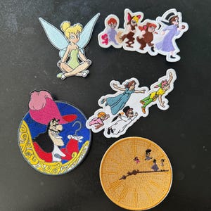 May include: A collection of five embroidered patches featuring characters and scenes from Peter Pan. The set includes Tinker Bell, the Darling children, Captain Hook in a circular frame, a flying group, and a circular clock face with silhouettes.