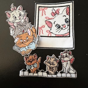 May include: A collection of embroidered patches featuring cartoon kittens. One patch shows three stacked kittens, another is a square polaroid-style frame with a kitten, and the third is a horizontal strip of three kittens sitting on piano keys.