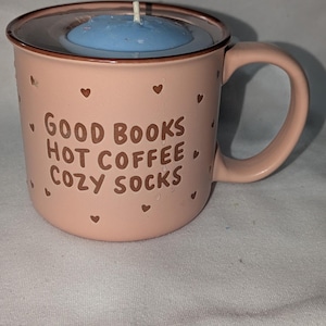 Puede incluir: Una taza de cerámica rosa claro con una vela azul en su interior. La taza tiene un asa y está decorada con pequeños corazones y las palabras "GOOD BOOKS HOT COFFEE COZY SOCKS" en letras marrones. La vela tiene una mecha blanca.
