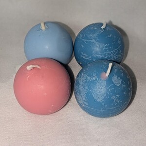 May include: Four spherical candles in shades of blue and pink. Two are a solid blue, one is pink, and the last two are a textured blue. Each candle has a white wick extending from the top. The candles are arranged on a white surface.