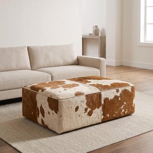 May include: A rectangular ottoman upholstered in genuine cowhide featuring a natural pattern of brown and white patches. The piece has a low-profile design with piped edges and a firm, boxy structure, showcasing the organic texture of the hide.