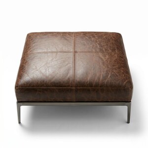 May include: A square, brown leather ottoman with a stitched, grid-like pattern. The ottoman has a dark metal frame and four short legs. The leather has a distressed appearance, adding to its rustic charm. The ottoman is a comfortable seating option.