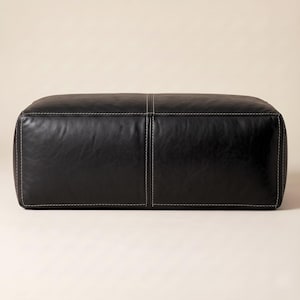 May include: A rectangular black leather ottoman featuring a smooth, slightly textured surface with visible grain. The piece is constructed with prominent white contrast stitching along the edges and a central vertical seam dividing the top surface.